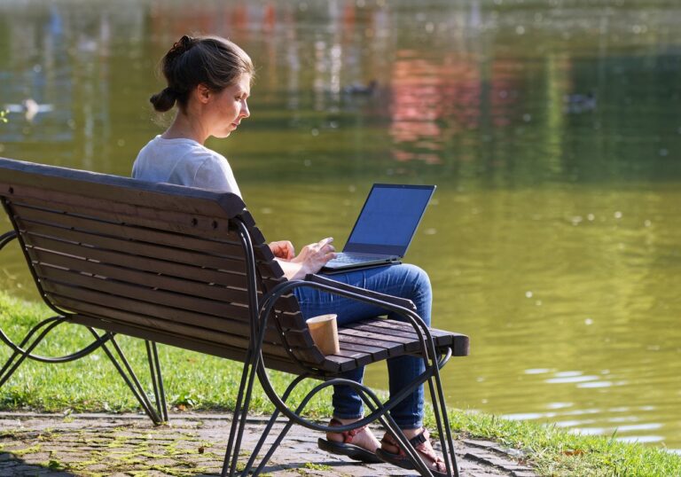 Woman working on a laptop on a park bench beside a lake